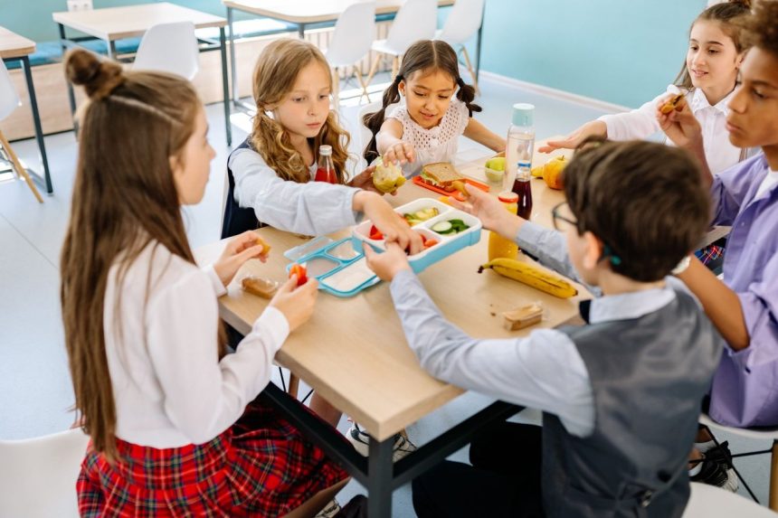 Grupo diverso de crianças faz uma refeição e compartilha lanches em uma cantina escolar.