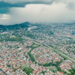 Vista aérea de uma área urbana extensa, com morros verdes ao fundo e uma tempestade se aproximando.