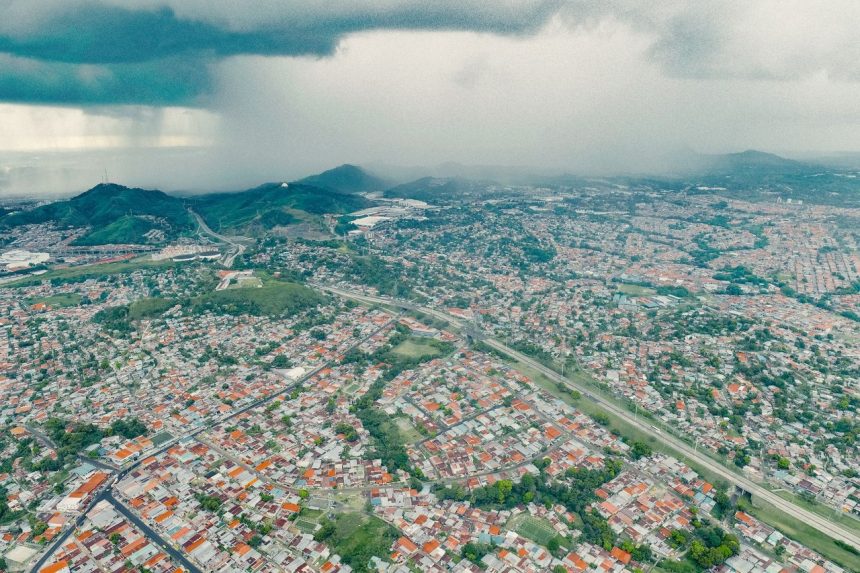 Vista aérea de uma área urbana extensa, com morros verdes ao fundo e uma tempestade se aproximando.