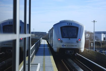 Trem elevado de conexão aeroportuária se aproxima de uma estação em um dia ensolarado.