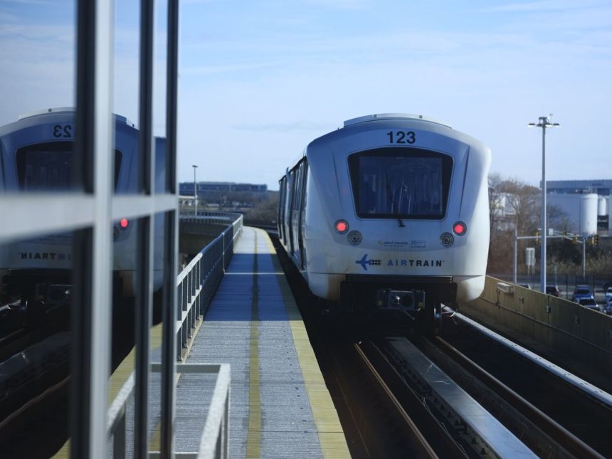 Trem elevado de conexão aeroportuária se aproxima de uma estação em um dia ensolarado.